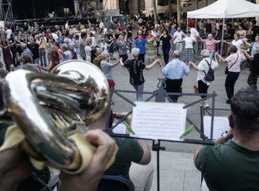 Gent ballant sardanes a la Plaça Vella acompanyats de la cobla, sobre l'escenari